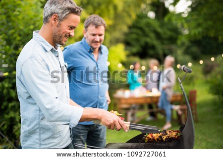 n a summer evening,  two men  in their forties prepares a barbecue for  friends gathered around a table in the garden