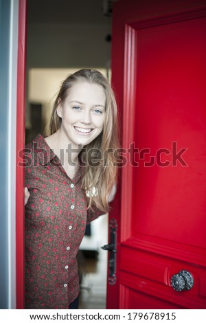 beautiful young woman opening a red door to welcome someone