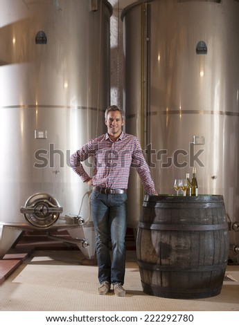 Professional winemaker looking at camera and tasting a glass of white wine in his cellar with modern stainless steel drums and traditional wooden barrels