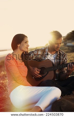 Romantic couple sitting on the beach at sunset with the man playing the guitar