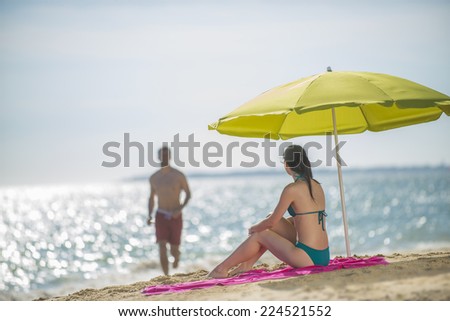 handsome couple in swimsuit at the beach the female sitting under a beach umbrella watches her man coming back from a sea bath