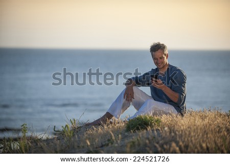 handsome man sitting on the beach at sunset and writing a sms on his phone