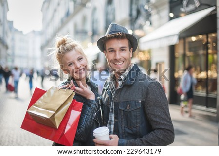 a trendy young couple walks in the city at christmastime, the young woman wears a leather jacket , shopping bags at her arm and the man a cup of coffee in hand