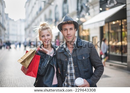a trendy young couple walks in the city in autumn, the young woman wears a leather jacket , shopping bags at her arm and the man a cup of coffee in hand