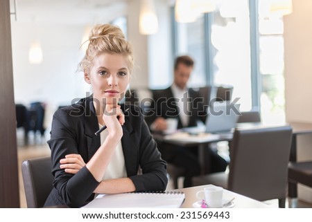 Portrait of a young businesswoman, sitting and writing at a table, a businessman blurred in the background