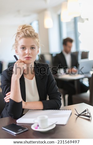 Portrait of a young businesswoman, sitting and writing at a table, a businessman blurred in the background