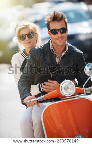 old fashionable couple riding  a vintage scooter in the street, man wears leather jacket and woman has a topknot and sunglasses