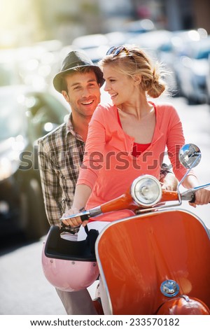 glamorous  young couple riding  a vintage scooter in the street, man wears a hat and woman has a topknot, sunglasses and a pink helmet