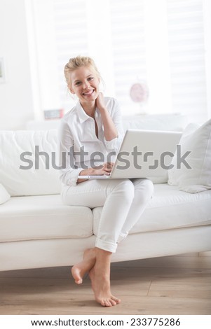 gorgeous blond woman barefoot sitting on a white couch at home and using a laptop