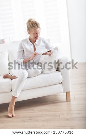 gorgeous blond woman barefoot sitting on a white couch at home and using a digital tablet