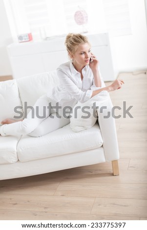 gorgeous blond woman barefoot sitting on a white couch at home and using her phone