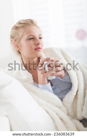 Beautiful blond woman holding a cup of tea lying on a couch, wrapped in a white blanket
