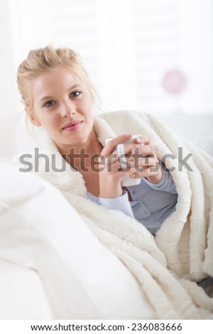Beautiful blond woman holding a cup of tea lying on a couch, wrapped in a white blanket