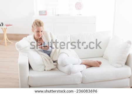 Beautiful blond woman using a digital tablet, lying on a couch wrapped in a white blanket