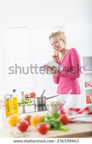 Cheerful young woman looking for a recipe on a digital tablet for cooking at home, vegetables on the work plan at foreground