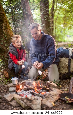in the evening a father and his young son roasting marshmallows on a campfire in the woods, their backpack  behind them