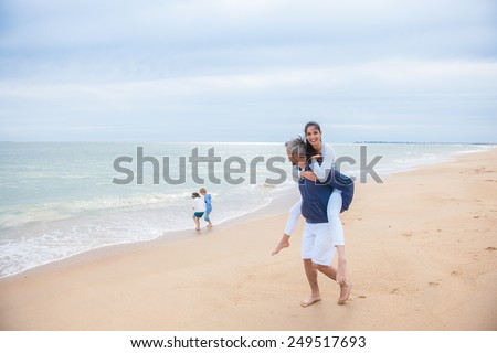 Family in casual clothes playing at the beach, dad is carrying mum while their children are playing in the sea waves