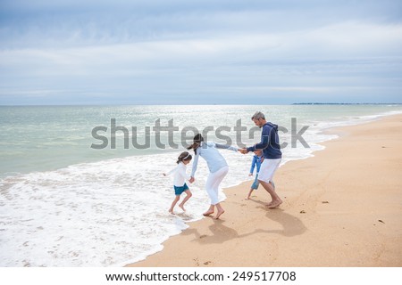 A four people family is playing in the sea waves, the parents and their two children are holding hands while running back from the sea waves