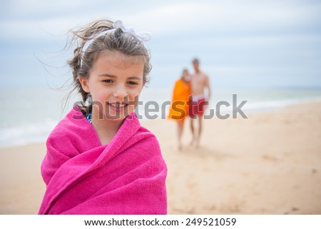 A smiling young girl look at the camera in her swimsuit, portrait of a six year old at the beach. She is wrapped in a pink towel, her parents are in the background holding each other.