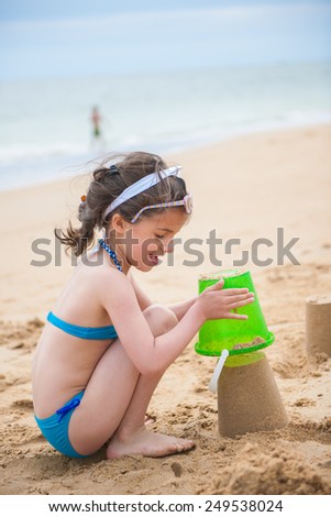 A six year old is playing in the sand with her bucket, a young girl is playing at the beach in her swimsuit.