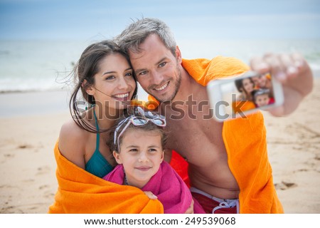 A young couple and their daughter are taking a selfie at the beach. Focus on the screen of the phone