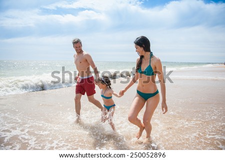 A six year old young girl at the beach is running with her parents in the sea waves, the family is in swimsuit