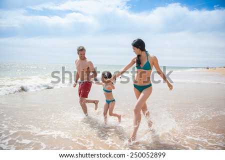 A six year old young girl at the beach is running with her parents in the sea waves, the family is in swimsuit