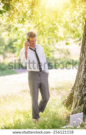 A good looking grey hair business man is standing naked feet in the grass, answering his phone.He is relaxing, his computer next to him, enjoying the shadow of the tree in a sunny day.