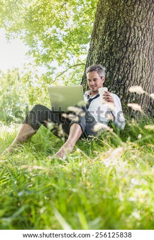 A nice looking grey hair business man is sitting against a tree in the grass, drinking coffee with his computer on his knees. He is relaxing, enjoying the shadow of the tree in a sunny day.