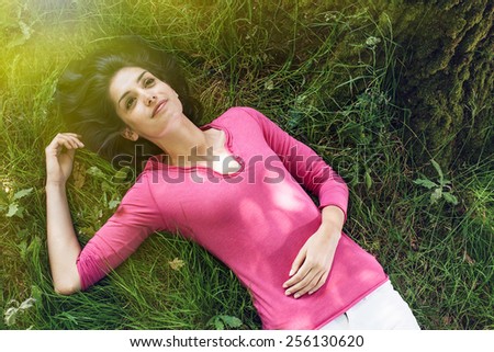 A beautiful woman is laying on her back in the grass, looking at the sky. She is relaxing, enjoying the shadow of the tree in a sunny day.