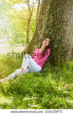 A nice looking woman is sitting against a tree in the grass, looking like she is dreaming awake. She is relaxing, enjoying the shadow of the tree in a sunny day.