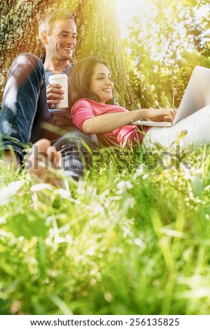 A nice grey hair man and a woman are sitting in the grass, looking at their computer. The man is sitting against a tree holding a coffee while his girlfriend is laying against him.