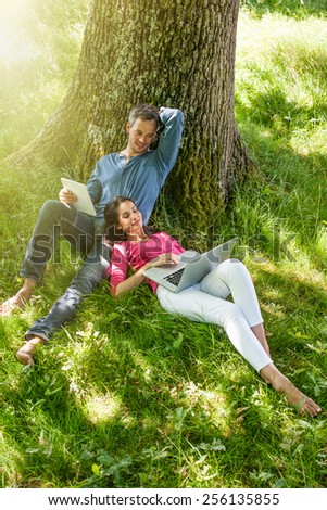 A nice grey hair man and a woman are sitting in the grass, looking at their tablet and computer. The man is sitting against a tree while his girlfriend is laying against him.