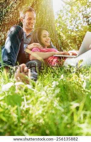 A nice grey hair man and a woman are sitting in the grass, looking at their computer. The man is sitting against a tree while his girlfriend is laying against him.
