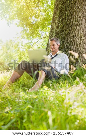 A nice looking grey hair man is sitting against a tree in the grass, looking at his computer. He is relaxing, enjoying the shadow of the tree in a sunny day.