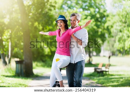 A lovely couple is having fun on a unicycle in the park. A beautiful woman is trying to ride the unicycle, while her grey hair boyfriend is holding her. They are wearing casual clothes.