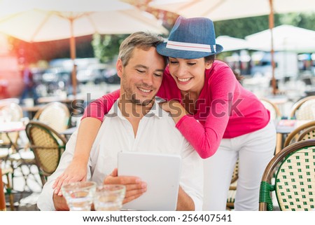 A smiling couple is enjoying drinks at an outside bar table in the city. The man is sitting at the table looking at a tablet and the woman is standing close to him.
