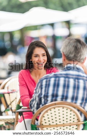 A loving couple is sitting at an outside bar table in the city. They are facing eachother. Focus on the woman. We can see the back of the grey hair man.
