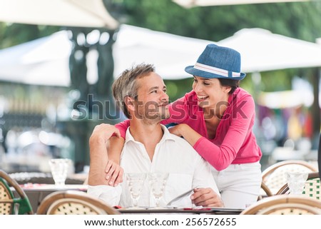 A smiling couple is posing for a picture at an outside bar table in the city. The man is sitting at the table and the woman is standing close to him. They are looking to each other in the eyes.