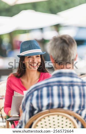 A loving couple is sitting at an outside bar table in the city. They are facing each other. Focus on the woman. We can see the back of the grey hair man.