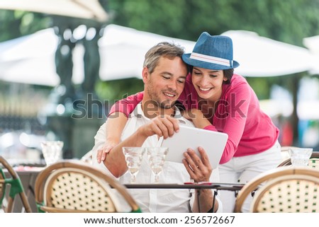 A smiling couple is enjoying drinks at an outside bar table in the city. The man is sitting at the table looking at a digital tablet and the woman is standing close to him.