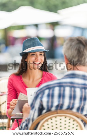A loving couple is sitting at an outside bar table in the city. They are facing each other. Focus on the woman. We can see the back of the grey hair man.