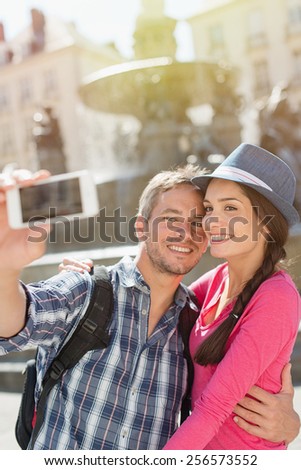 A loving couple is taking a selfie on their phone in the city . The woman is wearing   blue hat. The grey hair man is wearing a black backpack. They are looking at the camera with expressive faces.