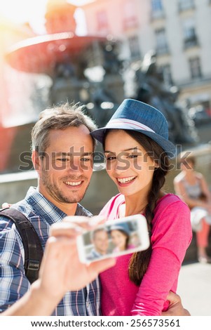 A loving couple is taking a selfie on their phone in the city . The woman is wearing   blue hat. The grey hair man is wearing a black backpack. They are looking at the camera with smiling faces.