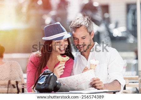 A couple is sitting at an outside bar table eating ice creams and looking at a map. The grey hair man with a beard is holding the map. The woman is wearing a blue hat, there is a camera on the table