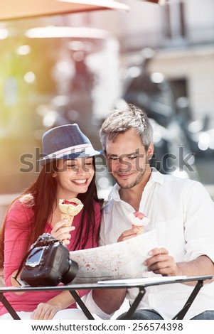 A couple is sitting at an outside bar table eating ice creams and looking at a map. The grey hair man with a beard is holding the map. The woman is wearing a blue hat, there is a camera on the table