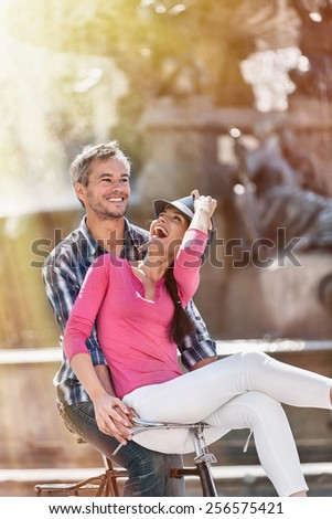 A smiling couple is riding a retro bike in the city center. The grey hair man is riding the bike while the woman is sitting on the handlebar. She is wearing a pink top and a blue hat.