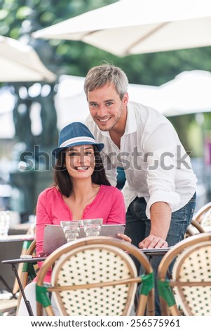 A smiling couple is posing for a picture at an outside bar table in the city. The woman is sitting at the table and the grey hair man is standing close to her.