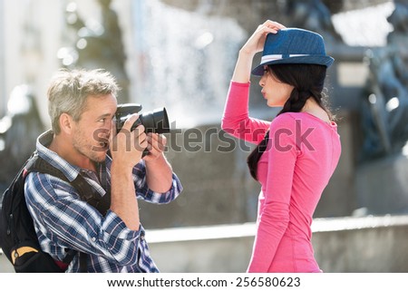 A loving couple is taking photos in the city center. The grey hair man with a beard and a black backpack is taking a closed picture of the smiling woman with a blue hat.