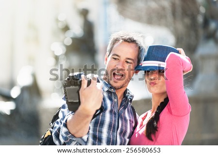 A young couple is taking a selfie on their camera in the city center. The woman is holding her blue hat. The grey hair man is wearing a  backpack. They are looking at the camera with expressive faces.